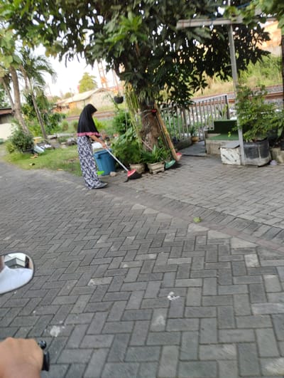 A person wearing a black hijab and a patterned skirt is sweeping a paved area. The person is standing next to a large blue trash can, holding a broom with a red broom head. Behind the person, there is a tree with lush green leaves and some smaller plants. To the right of the tree, there is a metal railing and more greenery. The ground is paved with interlocking gray bricks. In the background, there are some buildings and what appears to be train tracks. The scene suggests an outdoor environment, possibly a residential area or public space in Bringinbendo, Indonesia. The lighting indicates it is daytime, likely late afternoon or early evening due to the soft light. The overall atmosphere appears calm and focused on the task of cleaning.