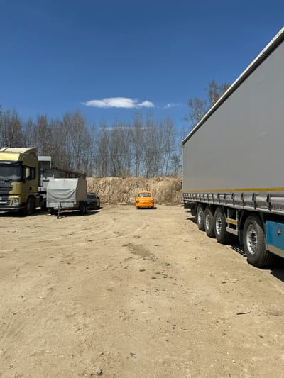 The image depicts a lot in what is presumed to be Sadovoe, Russia, due to the prompt. The ground is light brown, with sparse vegetation. Parked on the lot are a large semi-truck with a grey trailer, an orange microcar, a grey truck and a yellow MAN Xsan truck. The sky is blue, with scattered trees in the background.