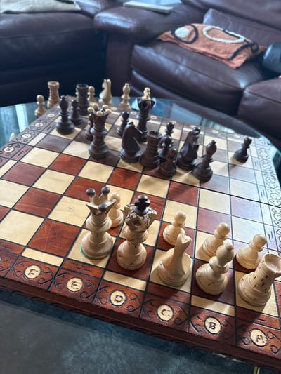 A slightly tilted, high-angle shot shows a chess set with wooden pieces on a board with brown and tan squares. The chess board has ornate, dark wood trim with letters carved into it. The pieces are mostly arranged in their starting positions. A brown leather couch with a decorative pillow is partially visible in the upper left. The surface beneath the chessboard appears to be reflective and dark. The location in Palaiseau, France is not discernible from the image.