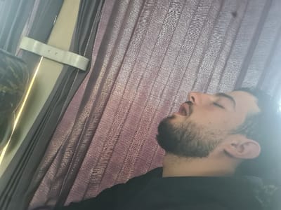 The image depicts a young adult male, appearing to be asleep, located in Al-Kiswa, Syria. The subject, positioned on the right side of the frame, has light skin, dark hair, and a short beard. His head is tilted back, with his eyes closed and mouth slightly open. He is wearing a dark-colored top. The background features layered curtains; a dark, solid-colored curtain is visible on the left, secured by a light-colored, flat strap with two visible perforations. Behind this, a lighter, textured, reddish-purple curtain is present. Bright illumination emanates from the left side, behind the curtains. A dark, patterned object is partially visible in the upper left background, suggesting it may be a headrest or part of a seat. The scene indicates an interior environment, possibly within a vehicle or a room with a window.