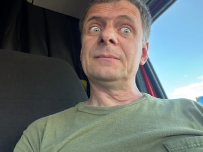 A close-up, selfie-style image shows a middle-aged man with wide, surprised eyes, a slightly open mouth, and light graying hair, wearing a faded olive-green t-shirt. He is seated in what appears to be a vehicle, with a dark fabric seat visible to his left and a red frame and bright blue sky to his right. The overall lighting suggests it is daytime, possibly outdoors. The image does not provide enough information to determine a specific location within North Platte, United States of America.