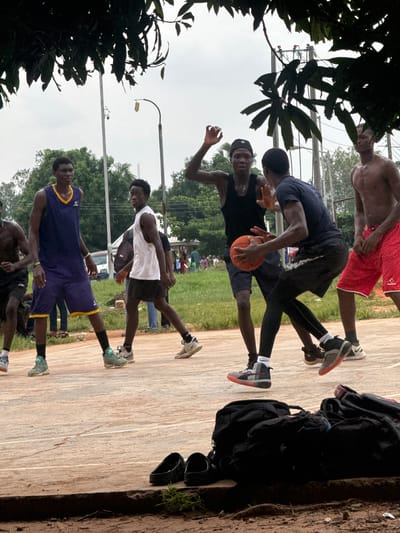 This is a medium shot, eye-level, outdoor photograph of a group of young Black men playing basketball on a dirt court in Amansee, Nigeria. The sky is overcast, and the light is diffused. The foreground is occupied by a pile of black duffel bags and a pair of black sandals. In the midground, several players are in motion. One player in a blue and yellow jersey is dribbling the ball. Another player in a black tank top is attempting to steal the ball. A third player in a white tank top and black shorts is running. A fourth player in red shorts and no shirt is standing on the right. The background shows trees, a street light, and a white van. The overall scene depicts an informal basketball game in a rural or semi-urban setting.
