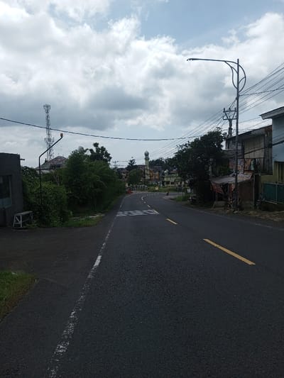 This is an eye-level photograph of a paved road in Gubbisamamidi, India, under a cloudy sky. The asphalt road features a white solid line on its left edge and broken yellow lines in the center, indicating two-way traffic. A pedestrian crossing is marked with white stripes further down the road.
On the left side, a dark, low-rise concrete structure with a visible window or opening is present, next to which a white, wooden-framed bench rests. Beyond this, dense green foliage lines the road, obscuring the view of buildings. A tall, lattice-structured communications tower is visible in the upper-left, behind a multi-story building with a reddish roof. An overhead street light fixture with an orange indicator light is also positioned on the left.
The right side of the road features utility poles with multiple overhead electrical or communication lines. A modern street light on a curved pole extends over the roadway. Alongside the road, a series of buildings are visible, including one with a small, covered roadside stall displaying various items. In the far distance, a prominent mosque with a minaret and dome stands among other structures. A single, small, dark vehicle, likely a motorcycle or scooter, is faintly discernible on the road near the distant buildings. No individuals are clearly visible.