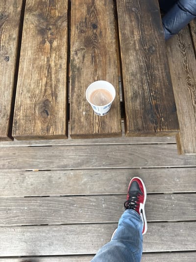The scene, located in Lièpvre, France, is depicted from an overhead perspective, showcasing an outdoor setting. In the lower frame, the left leg of an individual is visible, clad in light blue denim jeans and a red, black, and white sneaker. This leg is positioned on a ground surface composed of weathered horizontal wooden planks. Centrally, a rustic wooden table, constructed from wide, dark brown, visibly grained and knotted planks, holds a white disposable paper cup. The cup contains a light brown, foamy beverage and exhibits a faint blue design on its exterior. A portion of a dark blue or black puffy jacket, belonging to a second individual, is discernible in the upper right background, suggesting another person is seated at the table. The visible environment consists primarily of wooden structures.