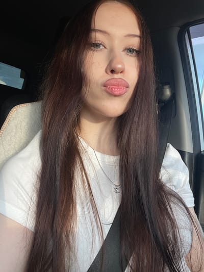 The media file is a portrait photograph taken in a car, with the subject's head and shoulders prominently featured. The subject is a young person with long, dark brown hair that extends across the frame. They have fair skin, light-colored eyes, and full lips with pink lipstick. A delicate silver necklace with a small pendant is visible around their neck. The subject is wearing a white t-shirt.

The setting appears to be inside a vehicle, suggested by the car's interior elements like the seatbelt and the textured material of the seat. The lighting suggests it is daytime, possibly with sunlight filtering in. The overall mood of the image is relaxed and casual, with the subject looking directly at the camera. There is no visible text in the image. The provided location context is Гавриловка, Russia.