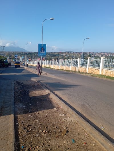 The image depicts a bright, sunny daytime scene along a road in Namanga, Kenya. The asphalt road, which appears to be on a gentle incline or decline, curves slightly to the right. On the left side of the road, there's a dusty, unkempt dirt shoulder scattered with rocks and litter, bordering a narrow paved sidewalk.
In the immediate foreground, a person dressed in a long, dark robe walks away from the camera on the road itself, heading towards the middle distance. Sunlight casts distinct, elongated shadows across the road and the dirt, suggesting either early morning or late afternoon.
Further down the road, other people are visible, along with vehicles, including a yellow three-wheeled tuk-tuk (or bajaj) and a larger, darker vehicle. A series of streetlights line the road. One prominent streetlight pole in the mid-ground holds a large, blue rectangular billboard. The billboard features a portrait of a man in a suit against a blue background. Below the portrait, clearly visible text reads "Hon. Mohamed Maalim Mohamud" and "Governor of Garissa County," with additional smaller text at the bottom.
On the right side of the road, a white and grey fence made of concrete pillars and metal railings separates the road from a lower-lying area. Beyond this, the background reveals a sprawling settlement of buildings and houses, characteristic of a town, spread across undulating terrain and hills. The sky above is clear and a vibrant blue, with a few faint clouds visible on the distant horizon. The overall scene portrays a bustling urban environment under clear weather conditions.