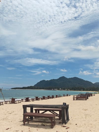 The image shows a beach scene in Monikeun, Indonesia. The foreground is dominated by a series of dark-stained wooden picnic tables set up on the sand. A mountain range is visible in the background, behind a line of trees and what appears to be small buildings or huts along the shoreline. The water is a light turquoise color, with gentle waves reaching the beach. The sky is predominantly blue with scattered white clouds. A numbered sign is affixed to the tabletop of one of the picnic tables. The sand appears relatively clean and light in color. There are no people visible in the image.