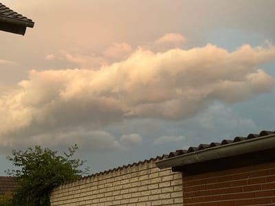 The image depicts a view of the sky above a brick wall and a roofline in Delbrück, Germany. The sky is filled with a mix of light and dark clouds. The clouds in the upper portion of the image are illuminated by warm, soft light, suggesting either sunrise or sunset. A large, elongated cloud formation with a golden hue dominates the central sky. The lower part of the sky transitions to darker, more ominous clouds, hinting at potential approaching weather.

In the foreground, a textured brick wall runs horizontally across the lower half of the image. A dark, wavy roofline, possibly made of corrugated material, runs along the top of the wall. To the left of the wall, a dense green bush or small tree is visible. The overall scene captures a moment in time characterized by changing atmospheric conditions and urban or suburban infrastructure. There are no people or animals visible in the frame. The lighting suggests it is either early morning or late evening.