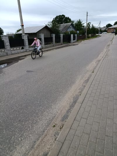 A person is riding a bicycle on a paved road in Смілавіцкі сельскі Савет, Belarus. The cyclist is wearing a light pink hooded sweatshirt and light-colored pants, along with a pink cap and white sneakers. They are carrying a backpack. The bicycle is black with blue accents on the wheels. The road is flanked by a paved sidewalk on the right and a fence and residential buildings on the left. The sky is overcast. The environment appears to be a suburban or rural area.