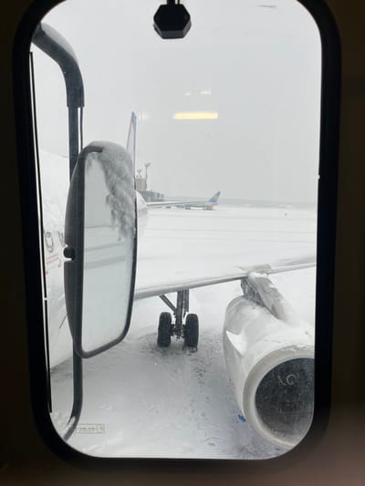 View from an aircraft cabin window shows a snow-covered tarmac and the wing of the plane, including its engine and landing gear. A partially snow-covered mirror is visible in the foreground. Another aircraft with a blue and yellow flag is further away. The scene is obscured by falling snow, and the window frame surrounds the view.