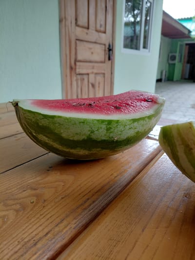 A sliced watermelon rests on a wooden surface, its vibrant pink flesh and green rind sharply contrasting with the warm tones of the wood. Another portion of the watermelon, also sliced, is visible in the upper part of the frame. The background depicts an outdoor scene with a light green wall, a wooden door with a window, and part of a building. The lighting suggests it is daytime. The context provided indicates the location is Akshi, Kazakhstan. There is no visible text in the image.