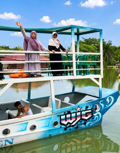The image depicts a two-tiered boat on a body of water, likely a lake or calm river. On the upper deck, two women are standing and posing for the camera. The woman on the left is wearing a lavender hijab and a matching long dress, and is waving. The woman on the right is wearing a patterned white and black hijab and a black dress. They are standing behind a white railing, with green chairs visible behind them. In the lower section of the boat, a young man wearing a white tank top is seated, looking down. The boat itself is blue with white and blue decorative waves painted on its side, and it has the word "WUG" emblazoned on it in stylized lettering. The surrounding environment includes lush green trees, palm trees, and a building with a red roof in the background. The sky is blue with scattered white clouds. The reflection of the boat and the surrounding scenery is visible on the water. The overall scene suggests a recreational outing on a sunny day. While the specific location is not visible in the image, the prompt mentions Eampat Kwalamencirim, Indonesia, implying this scene is set within that region.