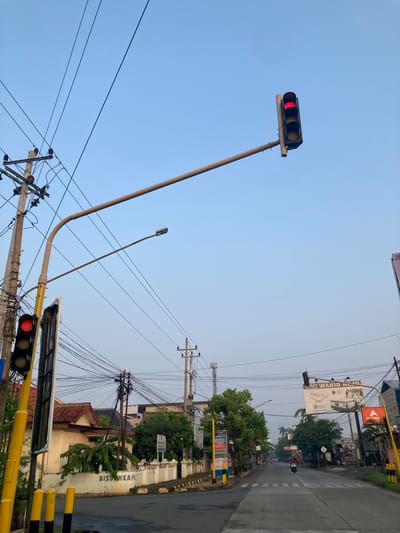 The image depicts a street scene in Kauman, Indonesia, under a clear sky. A traffic light with a red signal is prominent on the right side, positioned above a roadway. Utility poles with numerous crisscrossing wires are visible, indicating an urban environment with extensive infrastructure. The roadway shows a pedestrian crossing and appears to be asphalt. Buildings with tiled roofs and fences line the street, with some trees and vegetation interspersed. A motorcycle with a rider is proceeding down the road. Billboards are also present, suggesting commercial activity. The overall impression is a typical street view in a moderately developed area, possibly in the early morning or late afternoon given the lighting.
