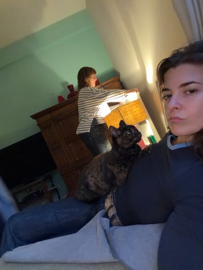 A selfie-style photograph features a woman with dark hair and eyes, wearing a dark blue shirt and jeans with a studded belt. A dark-colored, possibly brindle cat is perched on her lap, facing left. In the background, a woman with short brown hair is seen from the torso up, wearing a striped shirt and standing near a wooden dresser. Above the dresser is a lamp with a textured shade. The wall is light green. A dark rectangular shape resembling a television is partially visible below the dresser. The location is likely indoors. Specific details to locate in La Lucila, Argentina are not present.
