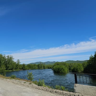 This is a landscape image taken in Apacha, Russia. The scene depicts a body of water, possibly a river, flowing alongside a lush green landscape. The water is a deep blue with ripples and reflections, suggesting a gentle current. On the right side of the water, a concrete embankment is visible, with a black metal structure, possibly a fence or railing, extending over the water. Beyond the water and embankment, dense green trees and bushes line the banks, with a mountain range visible in the distance. The mountains are covered in greenery, with patches of snow or light-colored rock appearing on their peaks. The sky is a bright, clear blue with some wispy white clouds stretching horizontally across it. The overall impression is one of a serene and natural environment. The time of day appears to be daytime, likely sunny, given the bright lighting and clear sky. There is no visible text in the image.