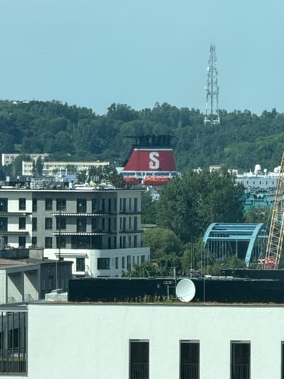 This is a vertical, wide-angle photograph taken from a high vantage point, looking down at a cityscape and a densely wooded hillside. The image is oriented such that the sky appears on the left and the ground on the right, suggesting the photo may have been rotated.

In the foreground, on the right, is a modern white building with multiple floors and dark-framed rectangular windows. Adjacent to this building is a dark, corrugated structure. Further to the left, a cluster of various buildings, some appearing residential and others more commercial, creates a dense urban environment. Many of these buildings have numerous dark windows.

Dominating the left and central portions of the image is a steep, heavily forested hillside. The trees are a lush green, indicating a warm season. Emerging from the hillside are several prominent structures. A large, red sign with a white “S” is clearly visible. Above and behind this sign, parts of what appear to be an amusement park ride with yellow scaffolding are visible. Further up the hillside, a tall communication tower or antenna array stands out against the sky. A blue, curved structure, possibly part of the amusement park, is also discernible.

A white satellite dish is mounted on a pole in front of the dark, corrugated structure and near the modern white building. The sky is a pale, clear blue, suggesting fair weather and likely daytime, possibly mid-morning or afternoon due to the lighting. No people are distinctly visible. The overall scene depicts a blend of urban development, natural landscape, and recreational facilities. There is no discernible text beyond the letter "S" on the red sign. The location context of Gdynia, Poland, doesn't provide specific visual cues within this image itself.