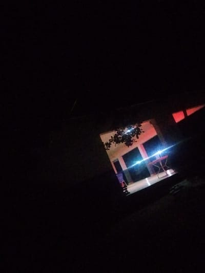 The image depicts a dimly lit scene, likely at night, in Mangu Town, Nigeria. Through an opening, a room or building interior is visible, illuminated by a mix of red and blue lights. The room appears to be a communal or recreational space, possibly a hall or a covered outdoor area. In the center of the illuminated space, a small table with a red top and metallic legs is present. To the left, a portion of a blue chair is visible. Above the table and chair, a cluster of dark-leaved foliage hangs, suggesting the presence of trees or bushes just outside the illuminated area. The overall lighting creates a dramatic and somewhat mysterious atmosphere, with bright lights creating lens flares and casting reflections on the floor. The darkness surrounding the illuminated opening implies it is late at night, and the ambient light suggests it is not raining. No people are clearly visible in the frame, and no text is discernable. The scene captures a quiet moment, possibly after an event or during a break in activity.