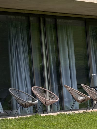 The scene depicts a building with a large window showcasing closed blinds, reflecting an exterior green environment. Outside, three contemporary chairs sit on a surface adjacent to green grass. The location and specifics of Chkalov, Armenia, cannot be determined from the image.