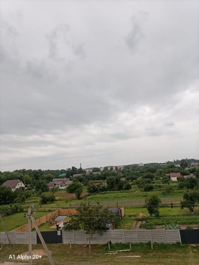 The image depicts a rural landscape under an overcast sky, likely in the Улукаўскі сельскі Савет, Belarus. The foreground features a gray concrete fence and a utility pole with wires. Behind the fence, a wooden fence encloses a grassy yard with a small structure, a barbeque grill, and a covered seating area. Further back, a field with crops is visible, and a person wearing an orange uniform is working in the field. Beyond the fields, a cluster of houses and trees form a village-like setting, with a tall red and white smokestack visible in the distance. The overall scene conveys a tranquil, everyday rural environment.