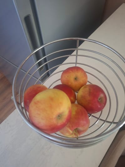 In Dumbrăvița, Romania, a metallic wire fruit basket is filled with approximately six red and yellow apples. The basket is positioned on a light-colored, textured surface, which appears to be a tabletop or counter. Behind the basket, a portion of a gray appliance, possibly a refrigerator, is visible. The background also shows a glimpse of a wooden floor. The lighting is ambient, suggesting an indoor setting.