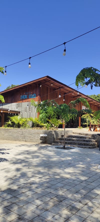 This is an eye-level, outdoor, daytime photograph of the exterior of a building with a wooden facade. The building is situated in what appears to be a public space, possibly a restaurant or cafe, with a paved courtyard in the foreground.

The building itself has a prominent sloping wooden roof with corrugated metal sections. Blue text spelling "LHOK GAGE" is visible on the wooden facade, along with smaller text above it that reads "KAMPUNG BAHARI". String lights with exposed bulbs are suspended across the scene, adding an aesthetic element.

The courtyard is paved with square, greyish tiles, and there are visible shadows from trees cast across the pavement. To the right of the frame, a set of stone steps leads up to an elevated area. Landscaping features include palm plants and a small, slender tree in the center of the paved area. In the background, through an open section of the building, tables and chairs are visible, suggesting an indoor seating area. A water fountain is mounted on a stone wall to the left of the steps. The sky above is clear and blue.

The scene is devoid of any people. The overall impression is of a well-maintained outdoor area designed for relaxation or dining.

The building is located in Lhokseumawe, Aceh, Indonesia, a city on the northern coast of Sumatra, known for its economic importance. The name "Kampung Bahari" translates to "Maritime Village," which could indicate a thematic element of the establishment, or its proximity to the sea.