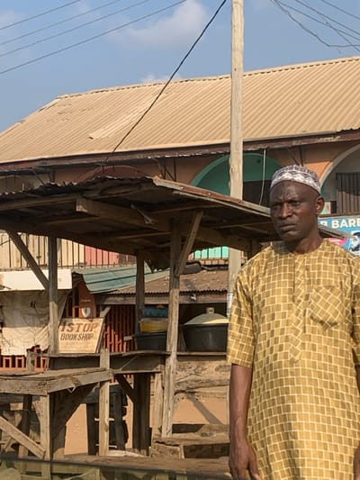 The scene depicts an outdoor commercial area in Nagazi, Nigeria. In the foreground, a dark-complexioned adult male stands facing forward, slightly right of center. He is bald, wears a white and grey patterned kufi, and a light brown/gold short-sleeved garment with a white grid pattern. His expression is neutral. To his left, a wooden market stall with a corrugated metal roof is visible. A white sign on the stall reads "ISTOP BOOK SHOP." Various containers, including black tubs, some filled with a white granular substance, are on or near the stall. A light-colored wooden utility pole stands vertically behind the man, extending to the top of the frame. In the background, a two-story building with a light brown corrugated metal roof is present. Portions of the building show arched architectural features, one painted light green/teal. A partially obscured sign in the background reads "R BARB," indicative of a barbershop. Multiple black utility lines traverse the clear, bright blue sky. The ground appears to be earth or dirt.