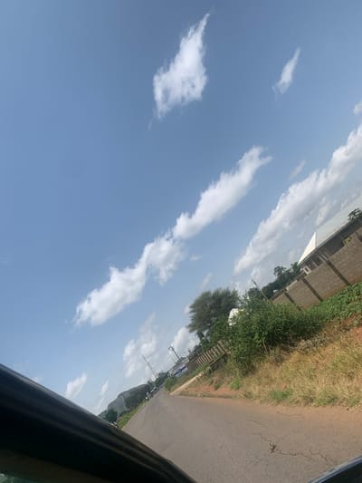 The image depicts a view from a vehicle traveling on a paved road in Kerang, Nigeria. The road curves to the right, leading towards a distant mountainous landscape. On the right side of the road, a low-lying building with a white roof is visible behind a concrete block wall. Dense green vegetation covers the area between the wall and the road, with dry grass and some smaller plants growing closer to the roadside. In the distance, construction cranes and tall poles are present, suggesting ongoing development. The sky is a clear blue with scattered white cumulus clouds. The foreground shows the dark interior of the vehicle, likely a car, obscuring the lower left portion of the scene.