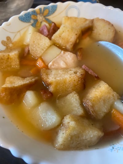 This is a close-up, overhead shot of a bowl of hearty soup. The soup appears to be a broth-based stew with chunks of potato, diced ham, sliced carrots, and croutons floating in it. The bowl itself is white with a decorative scalloped edge featuring a blue and orange floral pattern. A spoon is partially visible on the right side of the bowl, submerged in the liquid. The lighting is bright, suggesting an indoor setting. There is no indication of the city of Kolomna, Russia, or any people within the image.