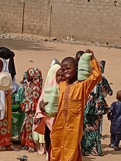 A group of African children, dressed in colorful traditional clothing, are gathered outside a brick building. One boy in the front, wearing an orange robe, carries a green bag on his head. Another child beside him holds a similar bag. Other children wear patterned fabrics and head coverings. The scene suggests a distribution of goods or food, with the children possibly waiting in line.
