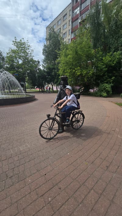 A young person wearing a white t-shirt, blue pants, and a light-colored hat sits on a bicycle. The bicycle is positioned in front of a bronze statue of a man wearing a hat. The statue is also seated on the bicycle, with its arms on the handlebars. The scene takes place outdoors on a paved area with red brick pavers. In the background, a fountain with multiple water jets is visible to the left, and a multi-story building is situated behind the trees. Several other people are seen walking in the background. The sky is cloudy. This location is in Ramenskoye, Russia.