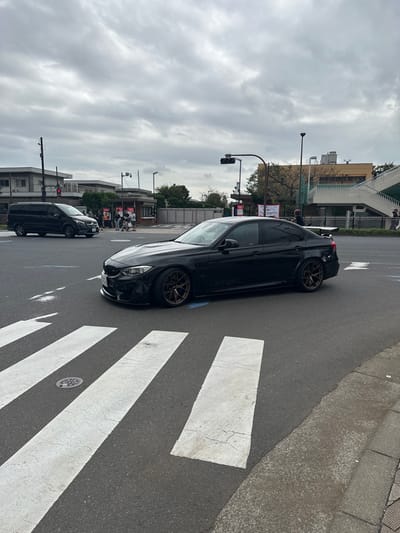 An exterior image captures an urban intersection in Shinagawa, Japan, under an overcast sky. A black, customized BMW M3 (F80 sedan) occupies the foreground and mid-ground, positioned on an asphalt road with its front wheels partially over white pedestrian crossing stripes. This vehicle features bronze aftermarket multi-spoke wheels, a lowered suspension, a front lip spoiler, and a large white aftermarket rear wing. In the left background, a black Mercedes-Benz V-Class van is visible on the road.

Multiple individuals are present in the background: a group stands near a low-rise building or booth on the left, and other figures are seen on an elevated pedestrian walkway with green railings to the right. A traffic light assembly with a visible red signal is positioned above the intersection. A white banner displaying "SHINAGAWA City Market" in red text is partially visible in the mid-ground on the right side. The environment includes power poles, streetlights, and various commercial or residential buildings. A circular manhole cover is embedded within the pedestrian crossing.