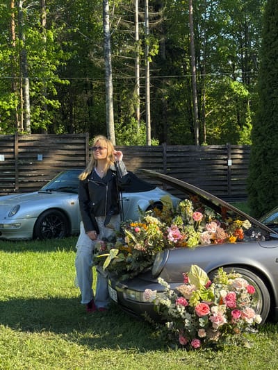 The image shows a young woman standing next to a silver Porsche car. The car's front is filled with an arrangement of flowers and greenery, extending from the open hood and encompassing the front tire. The woman is wearing a black leather jacket, a white top, light-colored pants, and sunglasses. She is touching her hair with her right hand. The background features a wooden fence, trees, and green grass. The scene is taking place outdoors on a sunny day. The license plate on the car indicates that the location is in Repino, Russia.