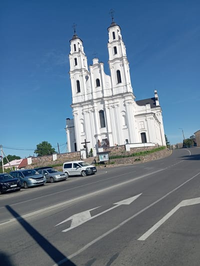 The image shows a white church with two prominent bell towers, characteristic of Baroque architecture, situated in the city of Hlybokaye, Belarus. The church is elevated on a stone wall embankment, with a paved road and several parked cars in the foreground. The sky is clear and blue. A white minivan and several cars, including a silver hatchback, a blue hatchback, and a black SUV, are parked along the roadside. The road features white directional arrows painted on its surface. The overall scene captures a typical day in this Belarusian city, with the majestic church serving as a central landmark.