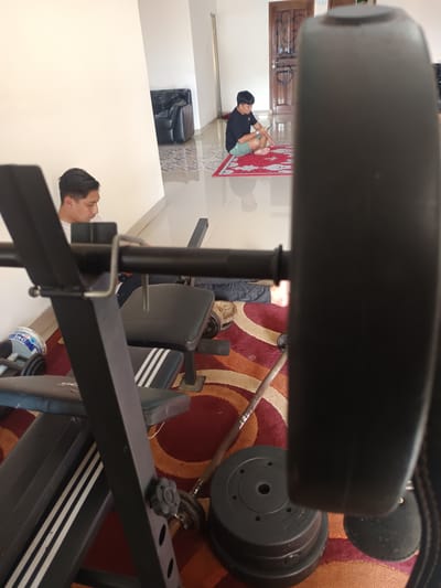 The image depicts a residential interior in Cianjur, Indonesia. A weightlifting bench press occupies the foreground. In the background, two individuals are present. One is kneeling on a prayer mat, using a mobile device, while the other is seated, looking towards the weightlifting equipment. The room's flooring consists of both tile and a red patterned rug, partially covered by weight plates and a barbell. A black leather sofa is visible in the background.