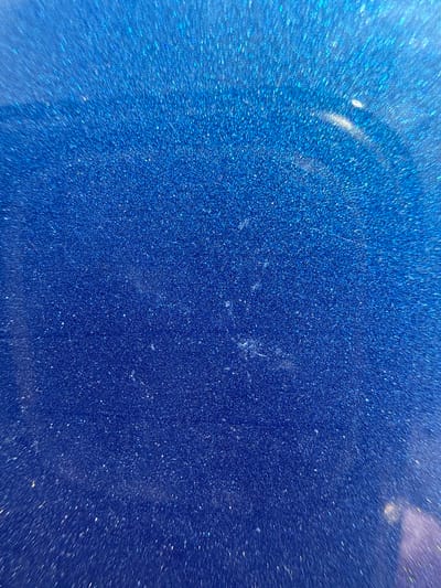 The image is a close-up, abstract shot of a sparkling blue surface. The surface is textured with numerous tiny, reflective particles that create a glittering effect, reminiscent of metallic paint or glitter. There are faint, irregular smudges or light reflections on the surface, particularly in the central and upper right portions of the image. The lighting suggests a smooth, possibly curved, material. There are no people, identifiable objects, or environmental features visible. This image is not locatable to a specific place in the city of Khmilna, Ukraine, or any other location, as it depicts a texture rather than a scene.
