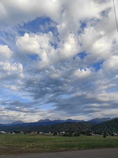 This media file captures a picturesque landscape scene in Dolan, Kazakhstan, under a dynamic sky. The image is dominated by a vast expanse of clouds, with patches of vibrant blue sky peeking through. The clouds vary in texture and shade, ranging from bright white, fluffy cumulus formations to darker, more brooding stratocumulus clouds, suggesting a day with changing weather.

In the mid-ground, a line of rolling green hills forms the base of a majestic mountain range. The mountains are imposing, with some peaks appearing snow-capped, hinting at a higher altitude and cooler temperatures even in what seems like a temperate season. Nestled at the foot of these hills is a small, dispersed village. The houses are varied in style and color, with rooftops in shades of red, white, and brown, and some appear to have fenced yards. A few scattered trees are visible among the houses, adding to the rural charm.

In the foreground, a stretch of lush green grass, likely a field or meadow, leads down to a dark, asphalt road. The presence of the road suggests human habitation and a connection to the wider world. The overall impression of the scene is one of natural beauty and rural tranquility. The lighting suggests it is likely late afternoon or early evening, given the soft quality of light on the clouds and mountains. There is no visible text within the image. The overall mood evoked is peaceful and awe-inspiring, showcasing the grandeur of nature and the quiet existence of human settlements within it.