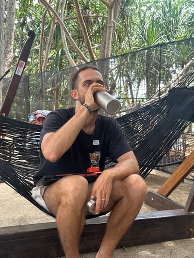 The image shows a man lying in a black hammock outdoors, surrounded by lush green foliage and trees. He is wearing a black t-shirt and appears to be drinking from a silver metallic bottle. His legs are crossed, and he has a ring on his left hand. The hammock is suspended from wooden beams, and the ground appears to be a textured surface, possibly concrete or stone. In the background, other people are visible in the distance, one of whom is holding a phone. The overall impression is that of a relaxed, casual setting, possibly a tropical vacation spot given the vegetation. The lighting suggests it is daytime, and the scene is likely outdoors in warm weather. There is a red and black object attached to the hammock near the man's leg, with some text on a white label attached to it, but the text is not legible. Another label with red text is visible on the wooden frame of the hammock, but this text is also too blurry to read.