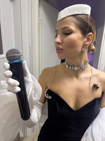 A woman stands in a doorway, holding a microphone. She wears a black velvet dress, a white pillbox hat, white gloves, a diamond necklace, and diamond earrings. A thin wire runs from her ear to her necklace. The background appears to be an interior hallway with white doors and walls. The location is specified as Истра, Russia.