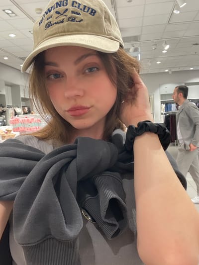 A young adult female is centered in the foreground, taking a selfie inside a brightly lit retail clothing store. She has light skin, medium brown hair, and wears a beige baseball cap with "ROWING CLUB EST. 1986 Riverside Park" embroidered in blue. Her attire includes a light grey ribbed top, with a dark grey sweatshirt draped over her shoulders. A black scrunchie is visible on her left wrist, and her left hand is raised near her temple, touching her hair. In the unfocused background, a modern retail environment is visible, featuring white ceiling tiles with recessed lighting, clothing racks displaying various garments (some in shades of pink and white), and display tables. To the right mid-ground, a male figure with short dark hair, wearing a light-colored jacket and trousers, is partially visible from the waist up, facing away from the camera and appearing to walk through the store. The scene is located in the city of Pompei, Italy.