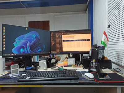 The image depicts a desk in an office environment, with two monitors displaying computer screens. On the left monitor, a Windows 11 wallpaper with blue and purple abstract shapes is visible. The right monitor shows an email client or a similar application with various text and data.

In front of the monitors, a black keyboard with Cyrillic lettering is positioned. To the right of the keyboard, a white coffee mug with a logo is present, along with a pair of eyeglasses and a stack of colorful sticky notes. A handheld scanner, likely a Zebra brand device, is placed in its charging cradle on the right side of the desk. Adjacent to the scanner is a small flag of Tajikistan. A white mouse sits on a black and red mousepad, and a smartphone is placed on the mousepad.

The background includes blinds on a window, and a portion of a door. The overall impression is that of a workspace, likely in an office building. There is no specific indication that this location is in the city of Yukhta, Russia.