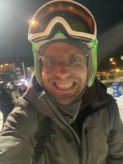 This image depicts a close-up, upward-angled selfie of an adult male in Jahorina, Bosnia and Herzegovina, at night. The subject is positioned centrally, visible from the chest up, and wears winter sports gear. He is equipped with a white and green ski helmet, over which orange-tinted ski goggles are positioned. Clear-framed eyeglasses are worn over his eyes. His face displays an intense, exaggerated grin, with his mouth open and teeth clenched, and his eyes wide and fixed on the camera. He wears a gray zippered winter jacket, with a dark interior layer visible, and a gray neck warmer or balaclava covers his lower face and neck. A dark strap, consistent with a backpack or bag, is visible over his left shoulder.
The background is dark, indicating nighttime, with various artificial light sources illuminating the scene. Snow is present on the ground. Blurred figures of other individuals are discernible in the left background, suggesting a populated area. One of these individuals appears to be holding a mobile device or camera. Distant buildings with illuminated windows and streetlights are visible in the mid-ground, indicating a developed area within a ski resort setting. A blue utility box or container is partially visible in the snow on the right side of the background.