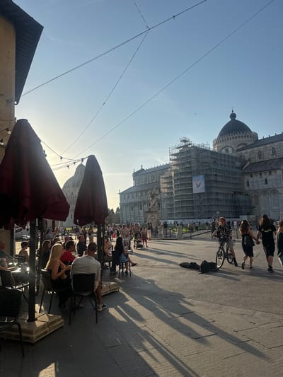 The image depicts a lively outdoor scene in what appears to be a historic public square, characteristic of Pisa, Italy. The setting is illuminated by the warm, low light of the late afternoon or early evening, casting long, distinct shadows across the light-colored paved ground.

In the background, grand architectural structures are prominent under a clear blue sky. A large, ornate dome, likely part of a baptistery or cathedral, rises majestically. Adjacent to it, another large building is partially obscured by extensive scaffolding, indicating ongoing restoration or maintenance work. Attached to this scaffolding is a prominent banner featuring the word "Peace" alongside a graphic of a dove, conveying a message of peace or solidarity. A small bird can be seen flying in the clear sky near the dome.

The foreground and midground are bustling with people. Many individuals are seated at outdoor tables under large, dark red umbrellas, suggesting an open-air cafe or restaurant. They appear to be socializing and enjoying the pleasant weather. Other people are walking along the paved area, and one person is visible on a bicycle. Overhead, several thin electrical or utility wires crisscross the sky. The overall scene captures a relaxed, communal atmosphere in a significant historical location.