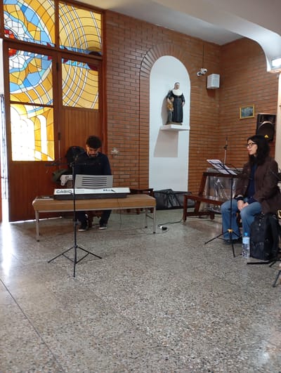 Two people are seated indoors, appearing to be in a church or a similar place of worship, given the stained-glass windows and the statue of a religious figure in an alcove. The person on the left is a young man with curly dark hair, wearing a navy blue sweater and dark pants, seated at a Casio keyboard. He is looking down at the keyboard, seemingly focused on playing. A microphone stand is positioned in front of him. On the right is a woman wearing glasses, a brown jacket, a navy blue top, and blue jeans. She is seated and looking to her right, with a music stand in front of her. Behind her, there is a speaker and a backpack. The floor is a terrazzo tile. The lighting suggests it is daytime, possibly late afternoon or early evening, given the way the light streams through the stained-glass windows, casting warm hues. The overall atmosphere seems calm and focused, suggesting a rehearsal or a quiet performance. The visible text is "CASIO" on the keyboard.