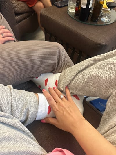 The image depicts an indoor scene within a private residence in Mitchells Plain, South Africa. The perspective is downward, focusing on an interaction between two individuals with a third visible in the background. In the foreground, a right hand with pink manicured nails and an oval-cut diamond ring on the fourth finger rests on another individual's sock-clad foot. The hand's owner is wearing light grey, textured casual trousers. The foot is covered by a white sock patterned with multiple red hearts, and its owner wears light brown, vertically ribbed casual trousers. In the mid-background, a brown upholstered ottoman or coffee table supports a circular mirrored tray. On the tray are a dark smartphone, a red wine bottle, multiple brown glass bottles, a clear beverage glass, and a wine glass containing red liquid. To the upper left, an upholstered armchair is partially visible, occupied by a third person whose lower legs are seen wearing an orange garment. The floor is light in color.