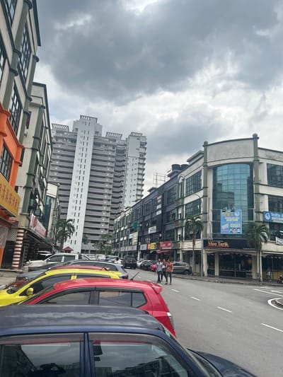 This image presents a vibrant, overcast street scene in an urban area, consistent with Subang Jaya City Council, Malaysia. The view is angled upwards from street level, encompassing a row of diverse buildings, a busy road, and a cloudy sky.
In the foreground and mid-ground, a variety of buildings line the street. To the left, a building with an orange and green roof features windows and some Chinese text, including "分店" (branch store). The central building is a multi-story structure with a contemporary design, including large glass windows on its upper floors. Its lower levels host businesses, with prominent signage for "TADIKA Golden Cradle" and its Chinese translation "金色摇篮幼儿园" (Golden Cradle Kindergarten), accompanied by phone numbers (018-2928965). Another sign advertises "RESTORAN WARISAN KAMPUNG" and "Mee Tarik," suggesting a noodle restaurant. Palm trees are scattered between the structures.
Further in the background, several tall, residential-style high-rise buildings with numerous balconies dominate the skyline. The sky is heavily overcast, filled with thick grey clouds, indicating a possibly humid or pre-rainy daytime weather condition.
The street itself is bustling with activity. Multiple cars of various colors, including red, yellow, and blue/grey, are visible, some parked along the curb, others potentially in motion. White lane markings are clearly visible on the asphalt road. Several pedestrians are scattered along the sidewalk and near the road, with two men notably walking in the mid-ground. The visible text in Malay, English, and Chinese languages reinforces the Malaysian setting.