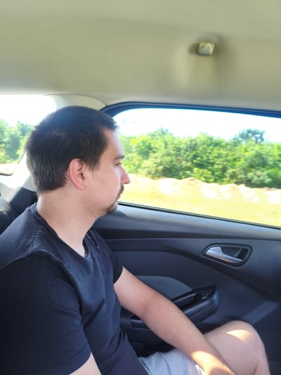 A medium shot captures a man from the chest up, seated in the back of a vehicle, looking out the window to his right. The man has short, dark brown hair and a short goatee. He is wearing a black t-shirt and grey shorts. The interior of the car is visible, including the dark grey door panel with a silver door handle and window controls. Outside the window, lush green trees and a bright yellow field are visible under a bright sky, suggesting a rural or suburban setting. The image appears to be taken during the daytime. The scene is not identifiable as being specifically in Baarle-Nassau, Netherlands, as there are no distinctive landmarks or visual cues to indicate this location.
