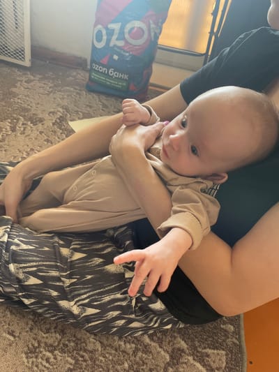 A medium shot captures a baby in a beige onesie being held by a person wearing a black top and patterned black and white pants. The baby is oriented towards the left of the frame, with its right hand extended outwards. An "ozon банк" bag in blue, red, and white is visible on the left side of the frame, resting on a floor that appears to be tiled or covered with a patterned rug. The location is Crokhovo, Russia.