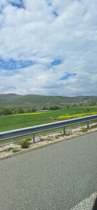 The image shows a roadside scene near Valkovo, Bulgaria. A gray asphalt road occupies the right side of the frame, with a metal guardrail separating it from a green field dotted with yellow flowers. Beyond the field is a hillside covered in green vegetation. The sky above is blue with scattered white clouds.