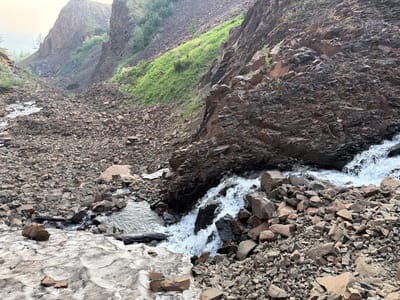 The image depicts a rocky, mountainous ravine with a small stream flowing through it. The ravine walls are steep and covered in loose rock and scree. Patches of green vegetation and small purple flowers are visible on the slopes. The stream is relatively shallow, with areas of both clear water and turbulent rapids. In the foreground, there is a patch of melting snow or ice. The overall environment is rugged and natural, suggesting a remote, undeveloped area. The image does not contain any people or specific landmarks that would directly place it within the city of Talnakh, Russia.