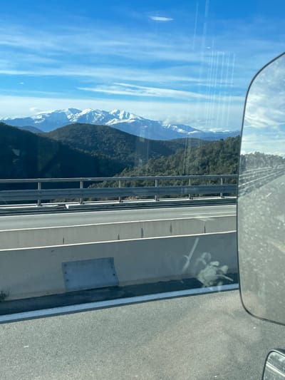 The image depicts a panoramic view from a moving vehicle on a multi-lane highway near Le Perthus, France, looking towards a mountainous landscape under a clear blue sky. In the immediate foreground, to the left, a light-colored concrete highway barrier is visible, featuring a rectangular metal plate bolted near its base. Above this barrier, a silver metal guardrail runs horizontally. The asphalt highway surface is marked with white lines, and reflections of vertical streaks and faint shapes are apparent on the window through which the photograph was taken. A significant portion of a vehicle's side mirror, likely a truck or bus, occupies the right side of the frame, reflecting a distorted view of the road and sky. Beyond the highway infrastructure, a valley with densely forested, dark green hills extends into the midground. In the far background, majestic snow-capped mountain ranges rise prominently, contrasting with the blue sky that contains sparse white clouds. No people or overt actions are visible; the scene captures a static landscape view from a dynamic perspective.